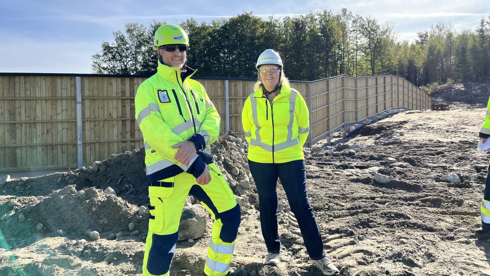 Christer Stenson, produktionschef på NCC, och Anneli Rossander, miljöspecialist på Trafikverket, står vid en byggarbetsplats med lerig mark och stängsel i bakgrunden under en solig dag.