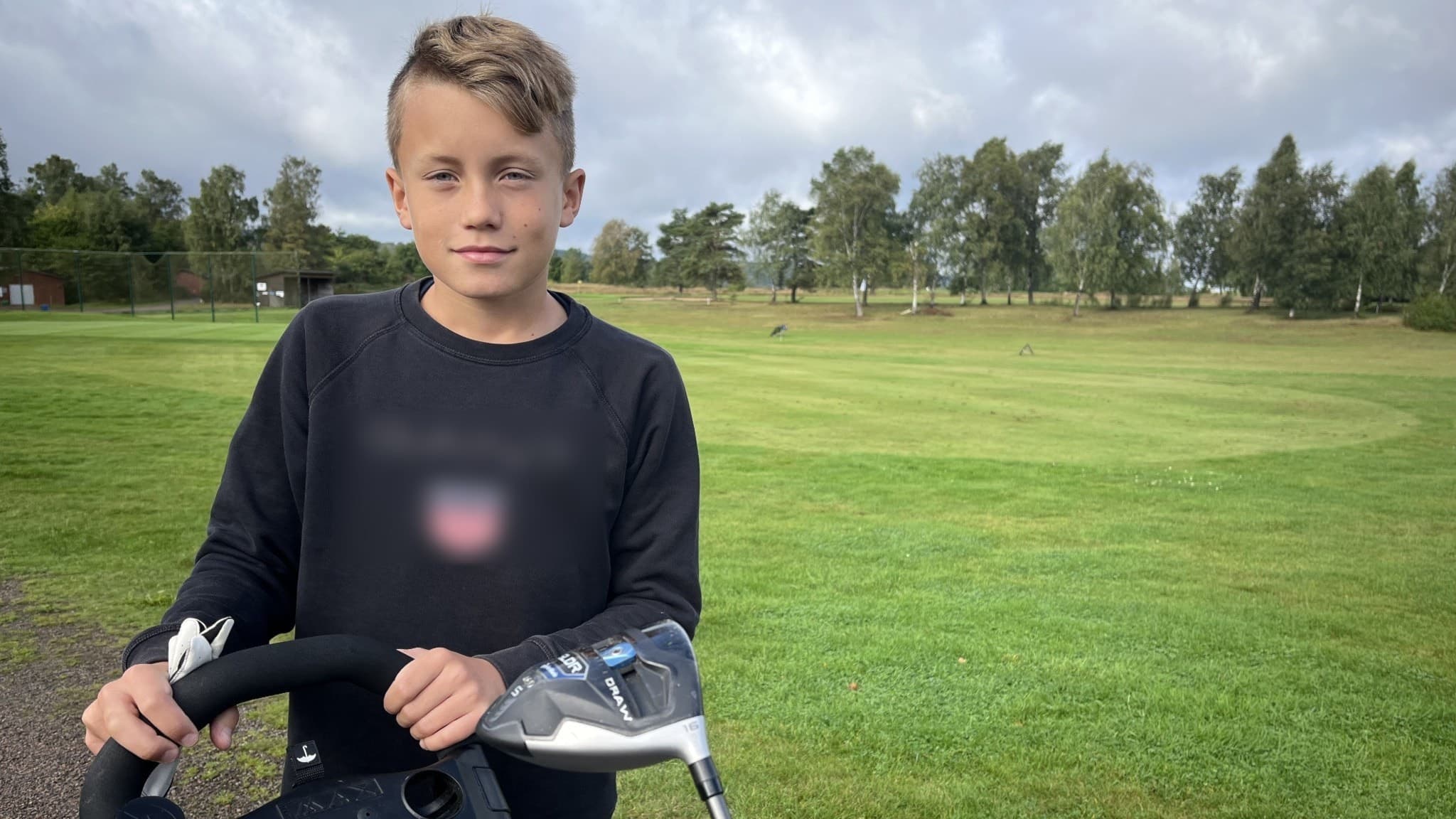 Samuel Olsson, 13 år, från Klippan står på en golfbana med en golfvagn och klubbor. Han har på sig en mörk tröja. Grön gräsmatta och träd syns i bakgrunden under molnig himmel.