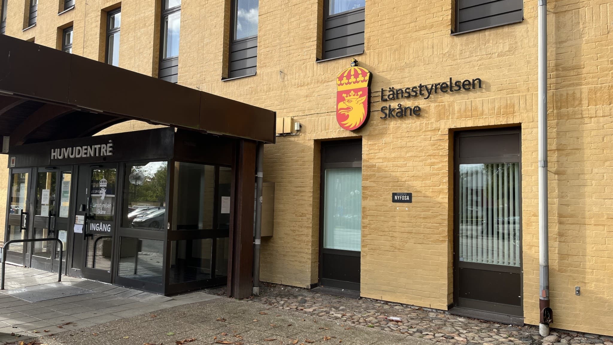 Entrén till Länsstyrelsen Skåne, en gul byggnad med stora fönster och Skånes emblem ovanför dörren. Området har stenläggning och glaspartier vid ingången.