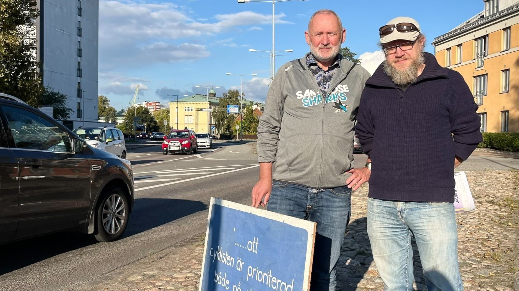 Tomas Stanislawski och en annan man står bredvid Långebrogatan med en skylt som de visar upp mot bilisterna som passerar. Bilar kör på vägen.