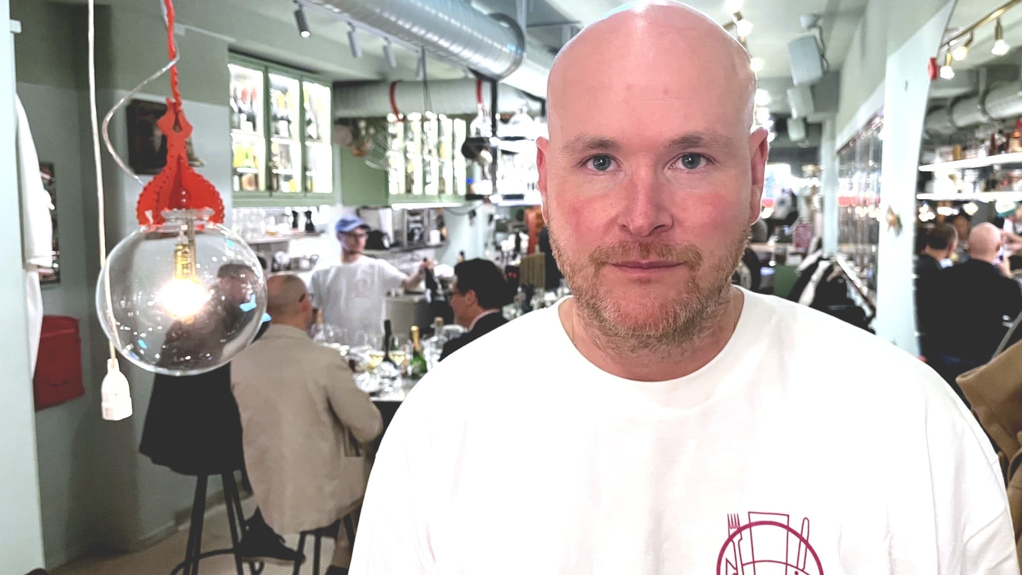 Johan Jureskog står i en modern restaurangmiljö med öppet kök i bakgrunden. Lokalen har industriell design med hängande lampor och hyllor fyllda med produkter.