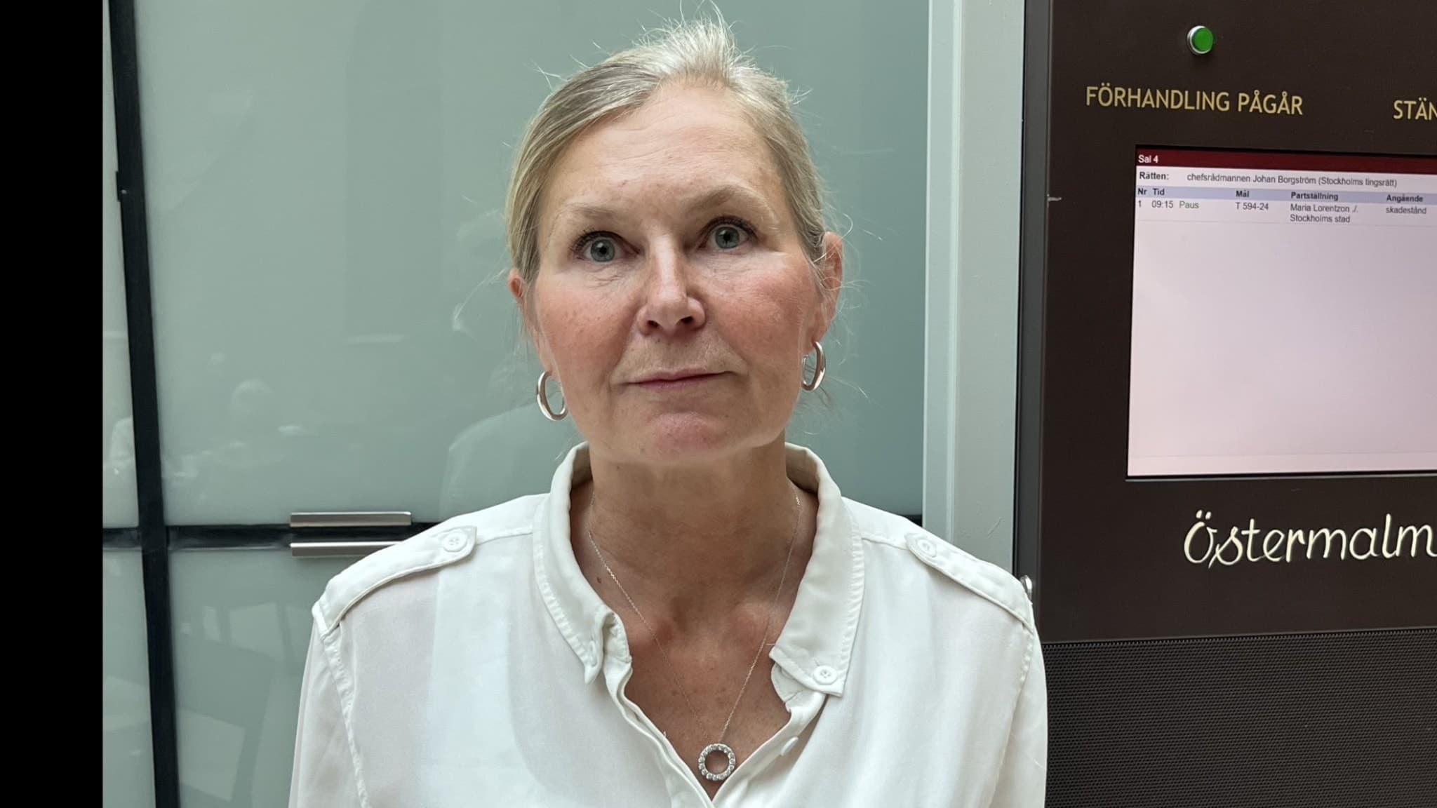Maria Lorentzon står framför en hissdörr med texten 'Östermalm' på en skylt. Hon bär en vit skjorta och har sitt hår uppsatt. Bilden är kopplad till nyheten om hennes son som tog sitt liv.