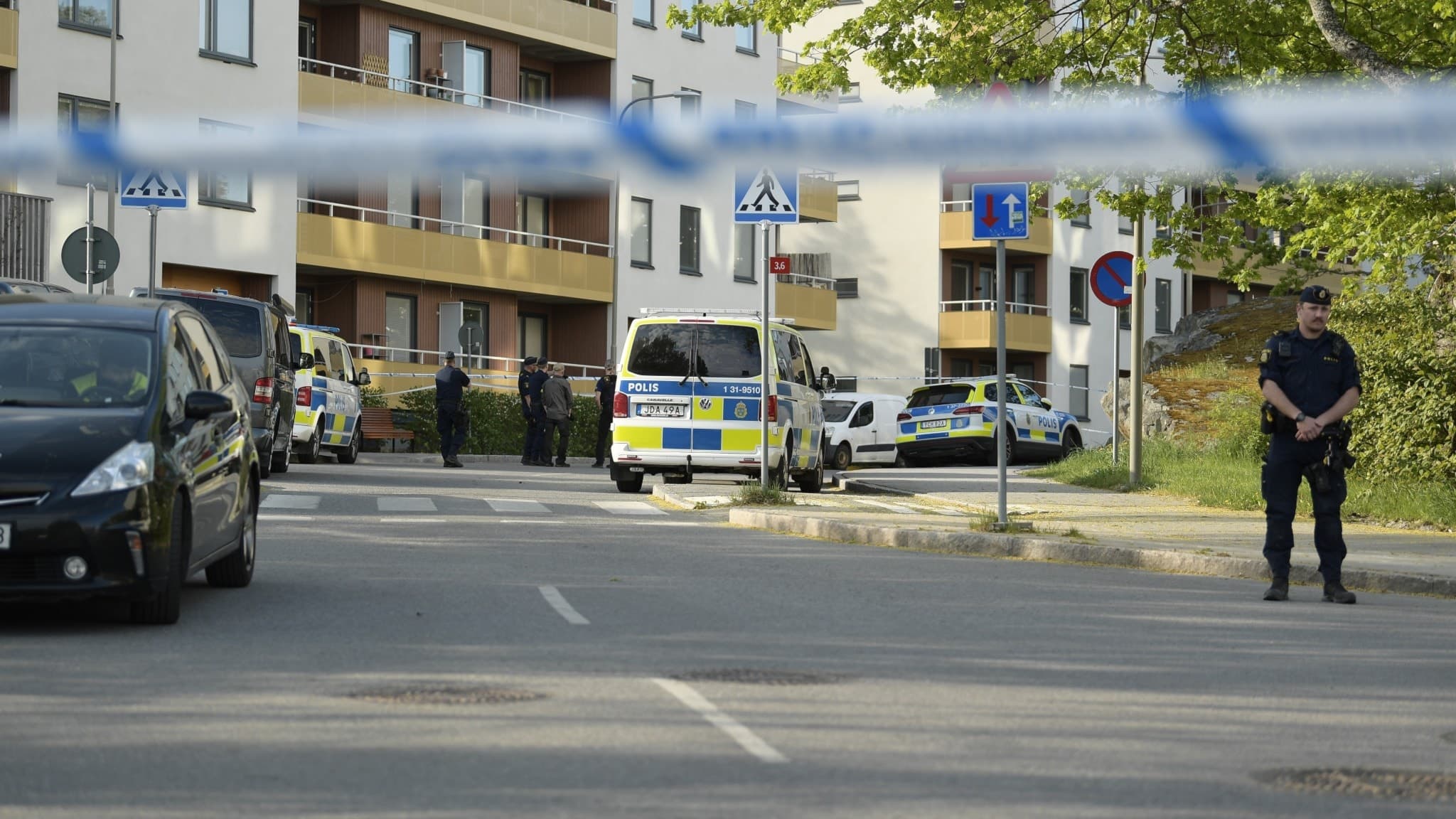 Polisavspärrningar vid en gata i Bredäng, Stockholm, efter en skjutning. Flera polisbilar och en uniformerad polis är på plats vid ett bostadsområde. Utredning pågår.