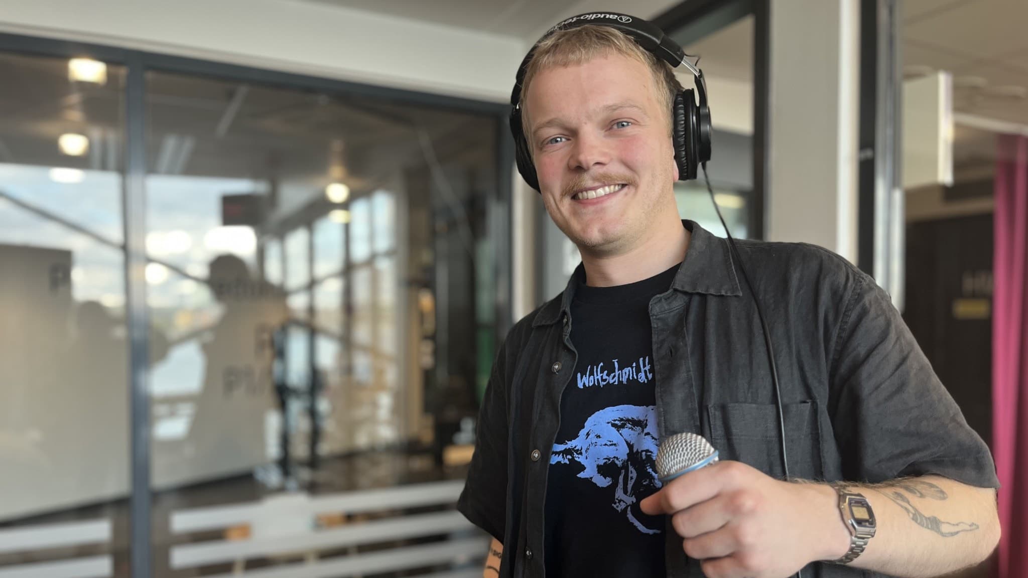 Filip Fernström håller en mikrofon och bär hörlurar i en modern studio med glasväggar. Han har på sig en svart skjorta och ett tryckt t-shirtmotiv, omgiven av kreativ studiomiljö.