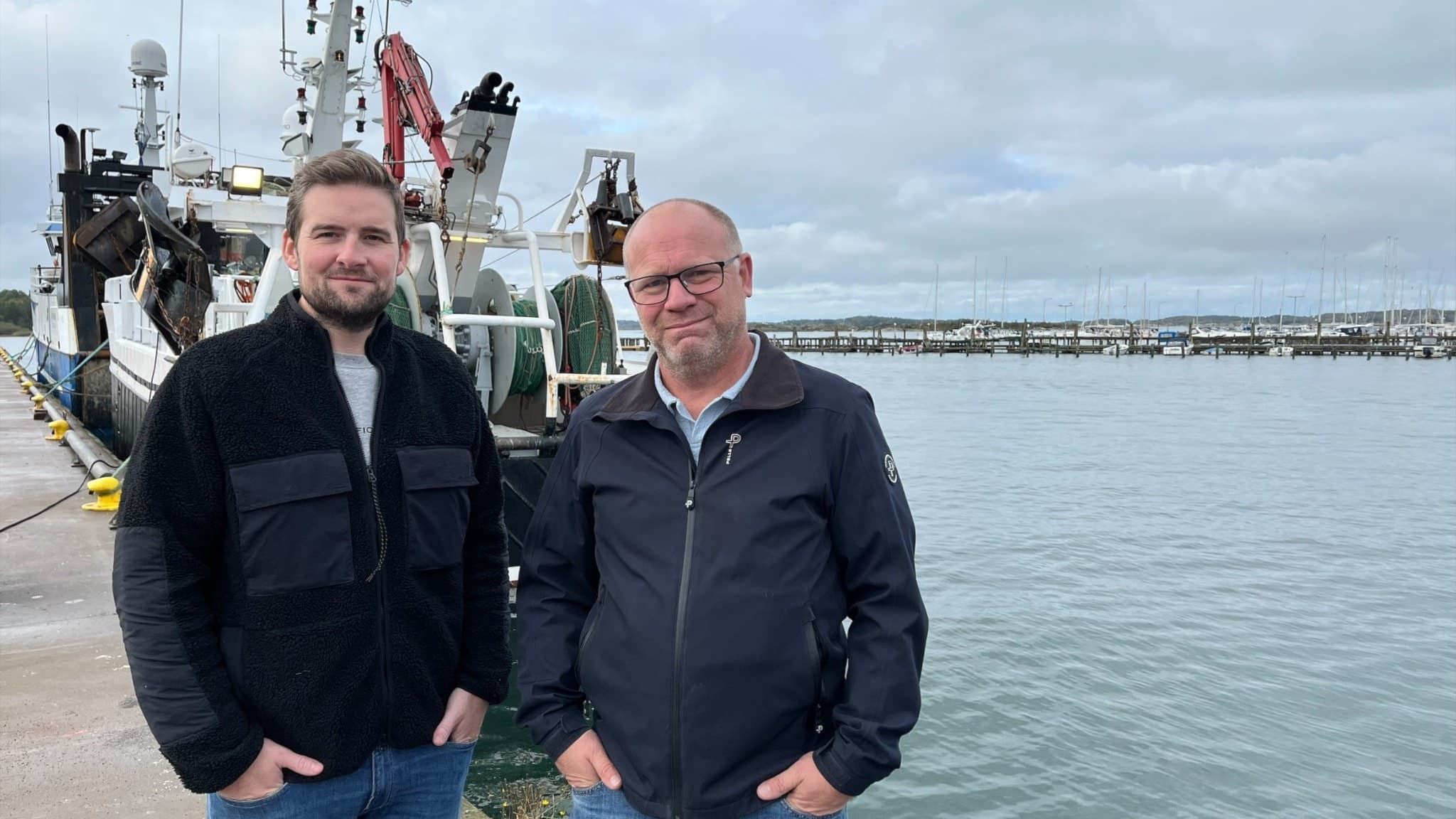 Johannes Claeson och Mathias Ivarsson står vid en hamn med fiskebåtar i bakgrunden. De är ordförande i den nya organisationen Vi Svenska Fiskare.