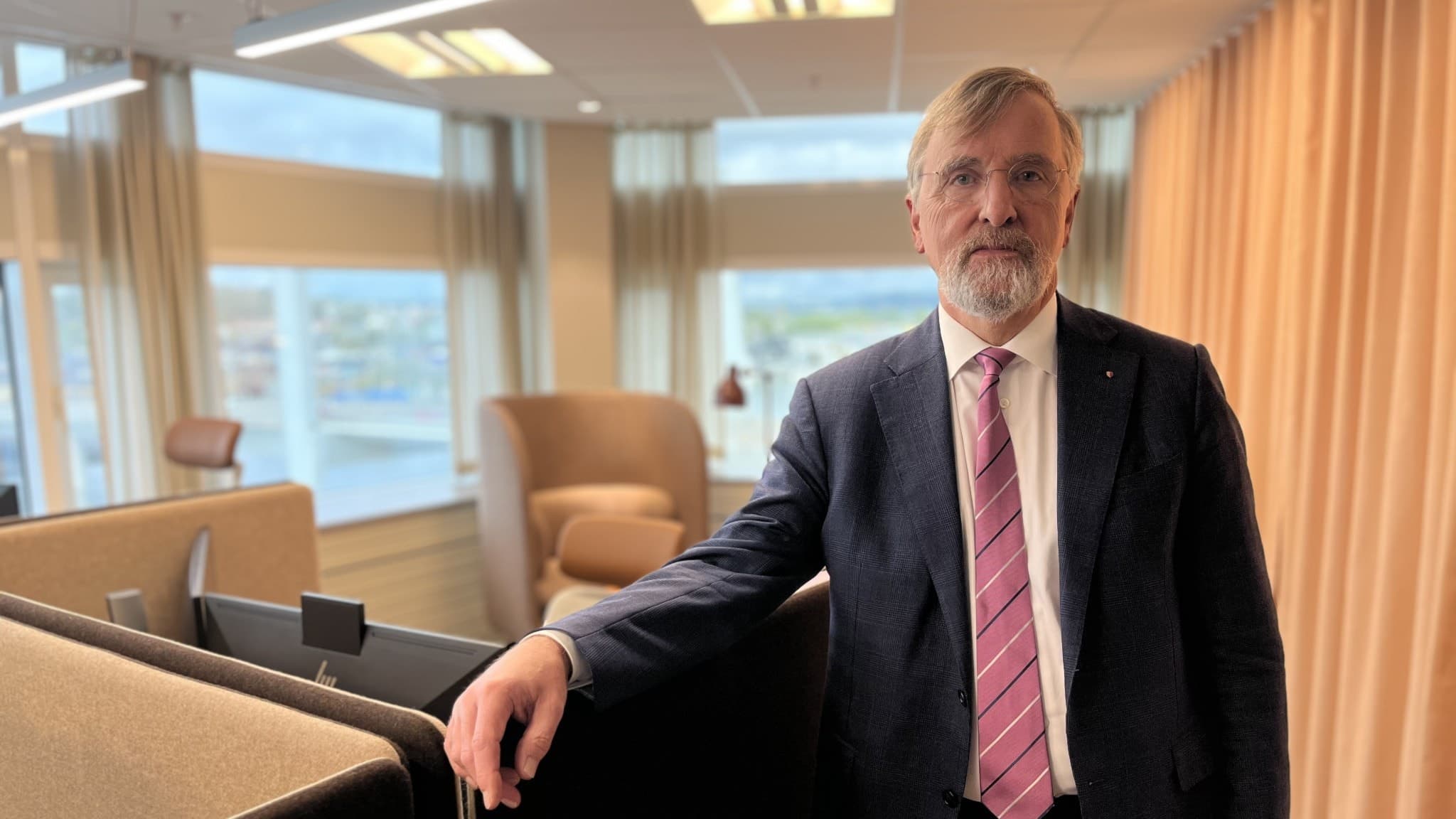En man i kostym med rosa slips står i ett modernt kontor med stora fönster som visar en stadsvy i bakgrunden. Ljusa möbler och gardiner skapar en professionell miljö.