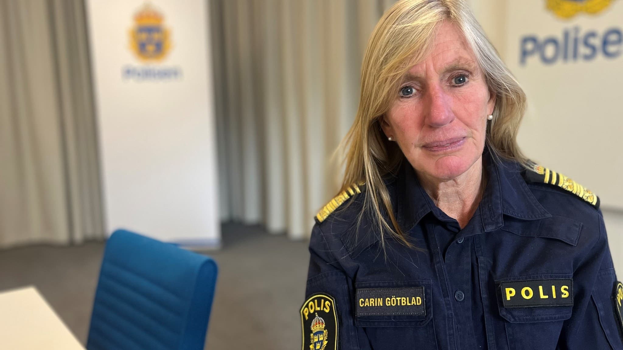 Polismästaren Carin Götblad, klädd i polisuniform, sitter i ett mötesrum med en vit bakgrund där polisens logotyp syns. 