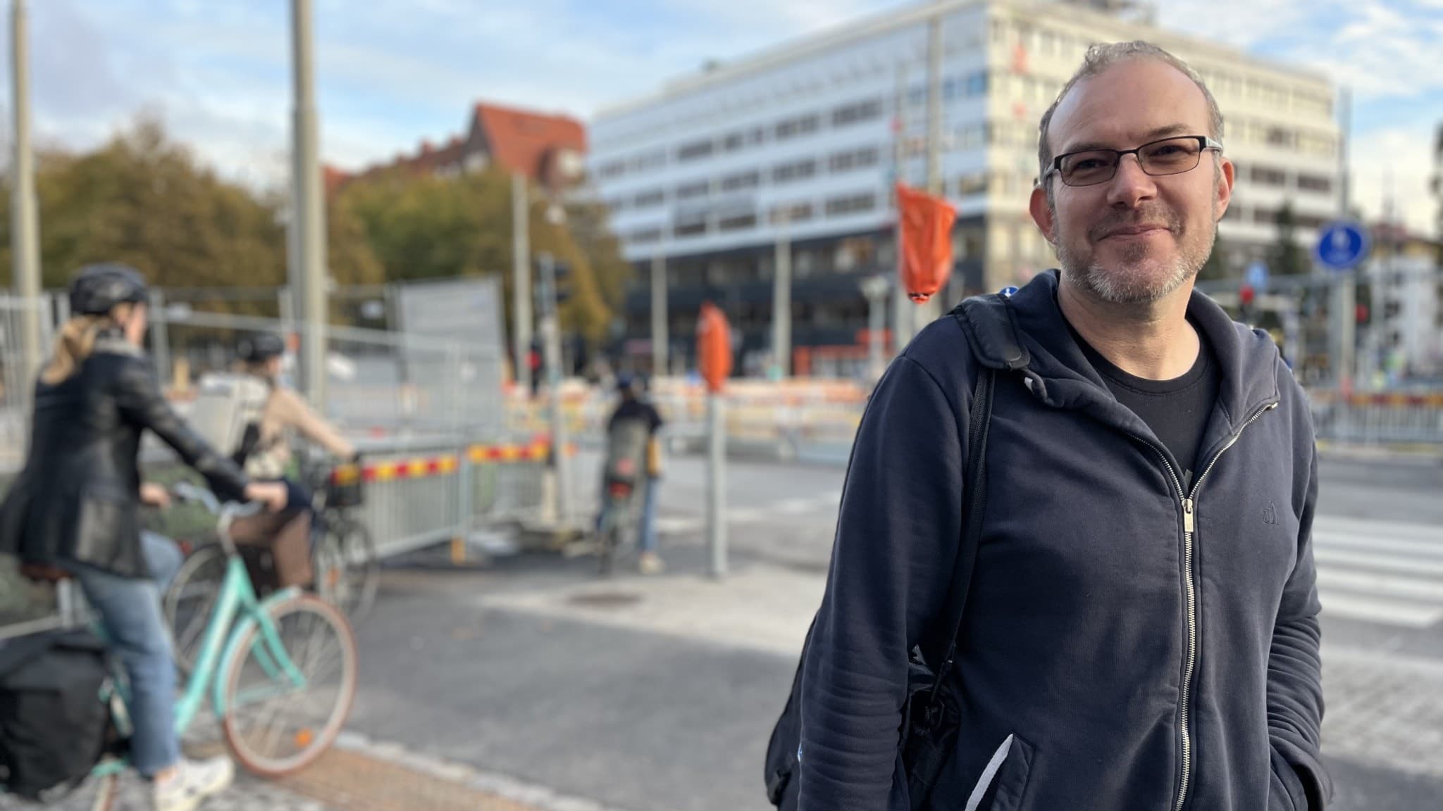 En person står vid en stadsgata med cyklister och fotgängare i bakgrunden. En modern byggnad och trafikinfrastruktur syns, inklusive avspärrningar och trafikskyltar. 