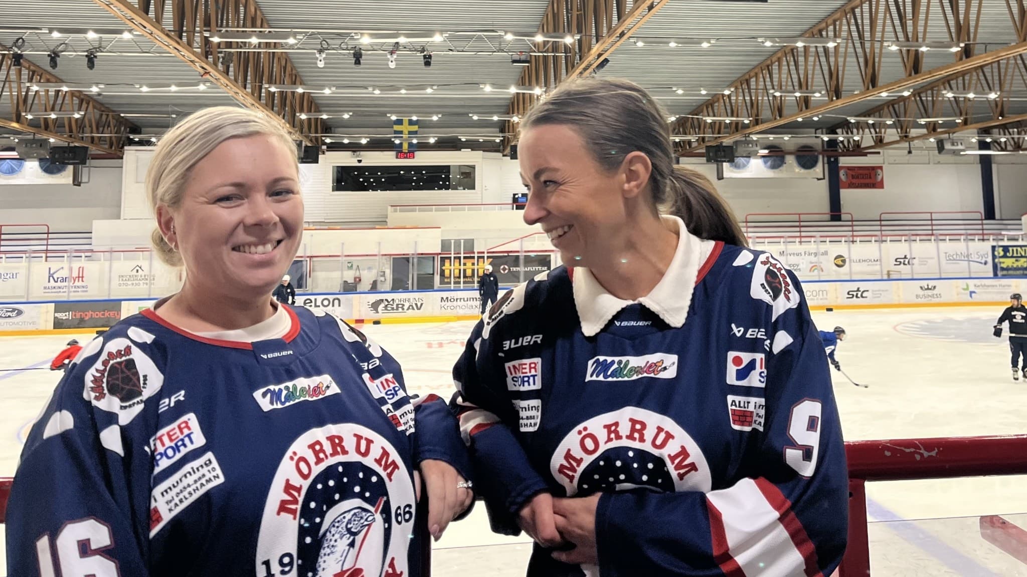 Emma Condrup och Maria Holmström, står vid sargen i en ishall iklädda Mörrum Hockey-tröjor. I bakgrunden syns en hockeyrink med spelare.