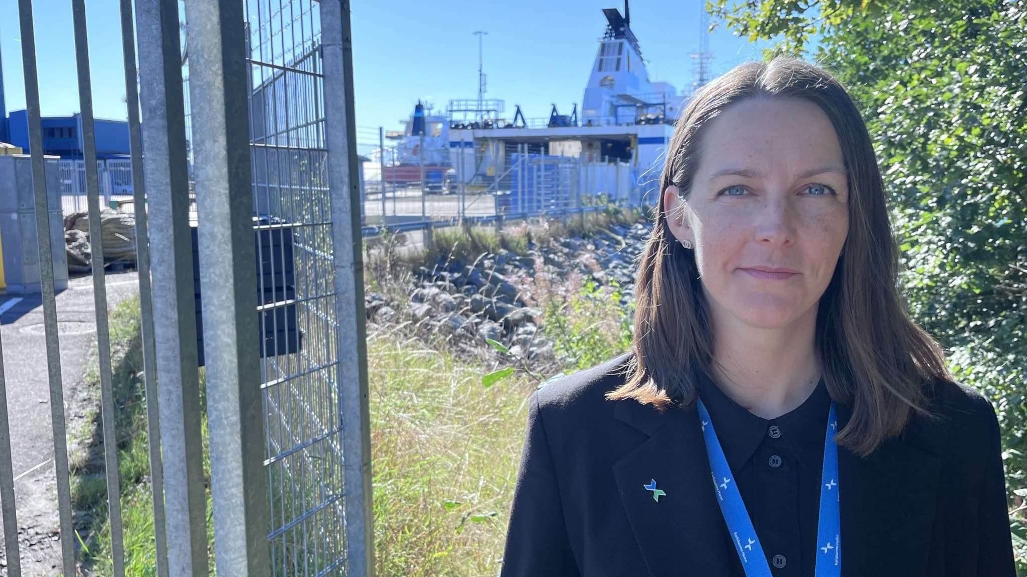 Caroline Säfström står framför ett stängsel vid en hamn med ett fartyg i bakgrunden. Hon bär en mörk kavaj och ett blått nyckelband runt halsen. Soligt väder och grönska i omgivningen.