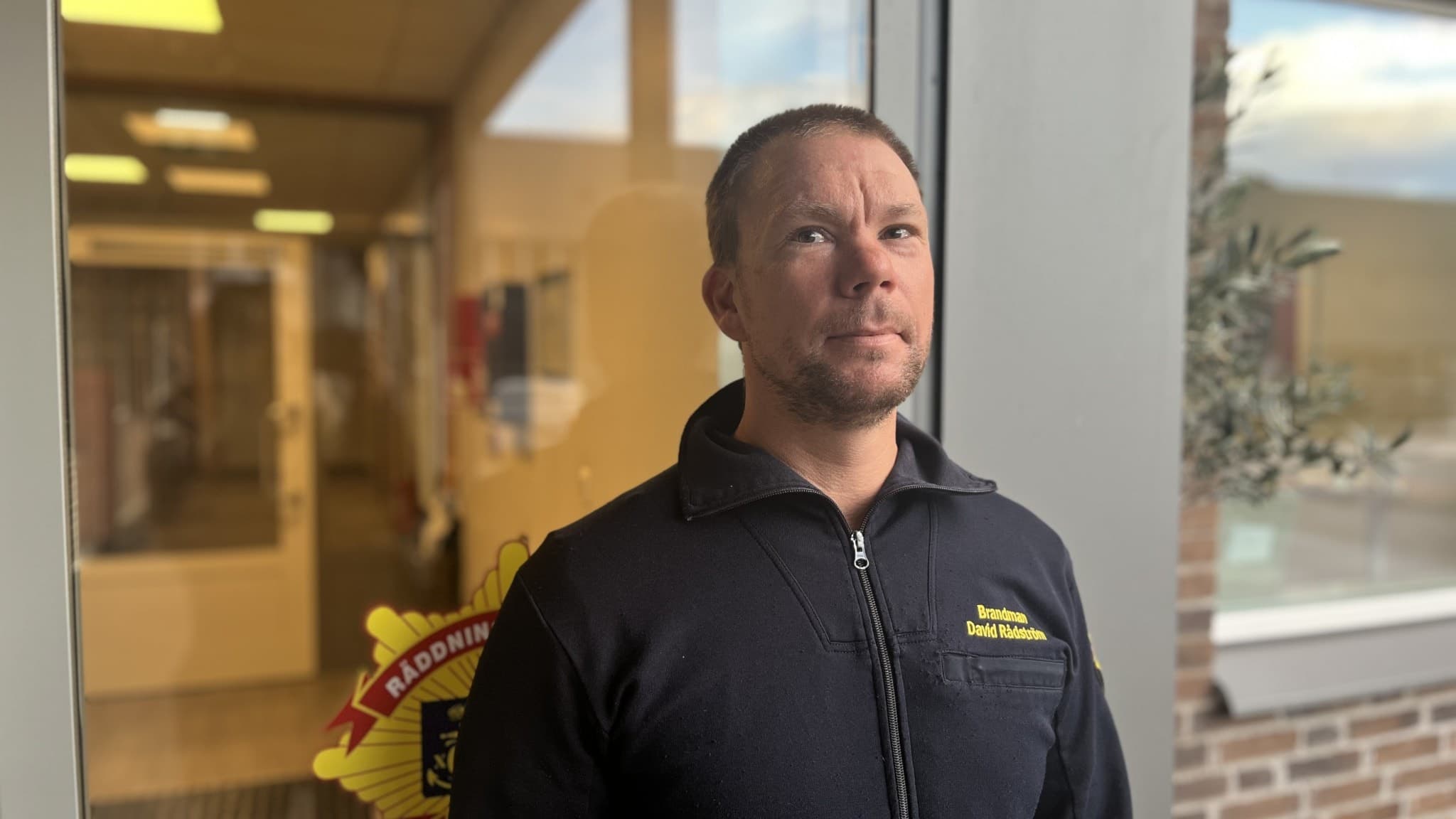 David Rådström står utanför en byggnad, klädd i en mörk tröja med texten 'Bring' på bröstet. I bakgrunden syns glasdörrar och en gul skylt.