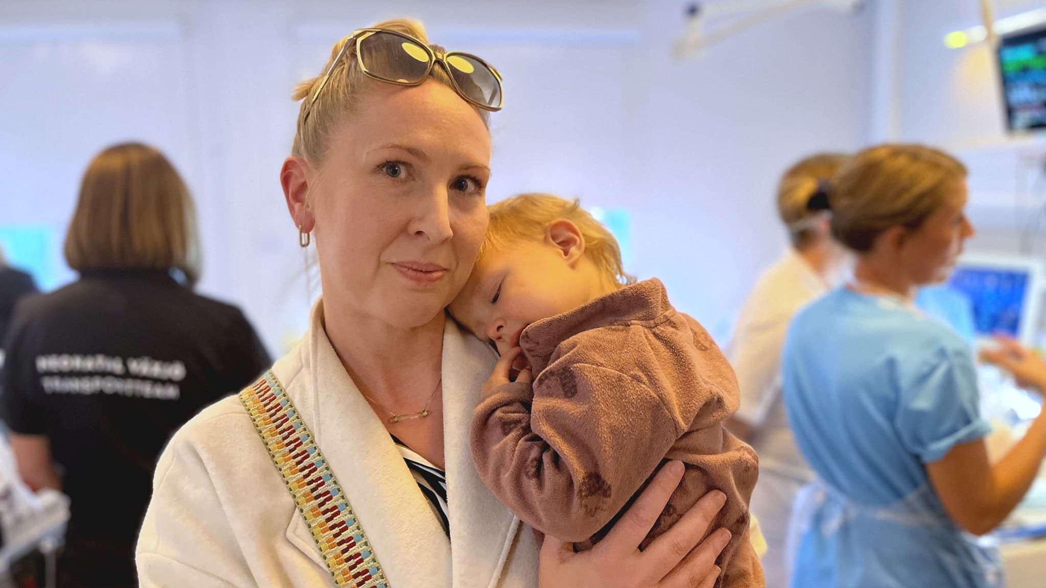 En kvinna håller ett litet barn i famnen på en vårdavdelning. Barnet har en brun tröja och lutar huvudet mot kvinnans axel. I bakgrunden syns sjukvårdspersonal och medicinsk utrustning.