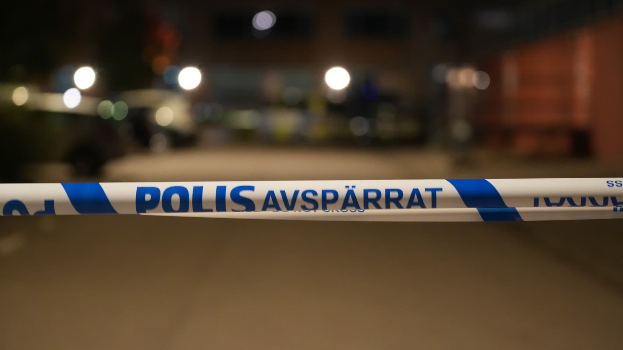 Ett område avspärrat med blåvit polisremsa, med texten 'Polis Avspärrat'. I bakgrunden syns suddiga ljuskällor och byggnader, vilket antyder en pågående utredning på platsen.
