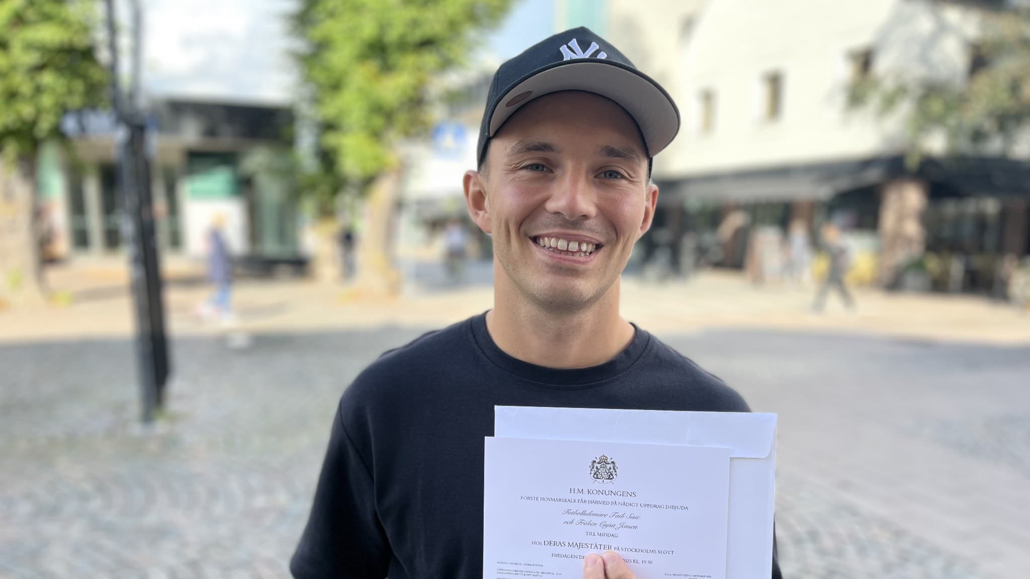 Fotbollsdomaren Taib Sisic håller i ett dokument med en kunglig inbjudan, stående utomhus på ett torg med byggnader och träd i bakgrunden.