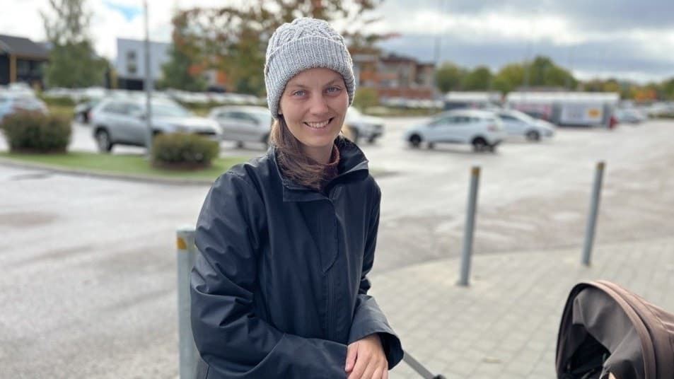 En person i mörk jacka och mössa står utomhus vid en parkeringsplats och håller i en barnvagn. Bakgrunden visar bilar, buskar och en mulen himmel.