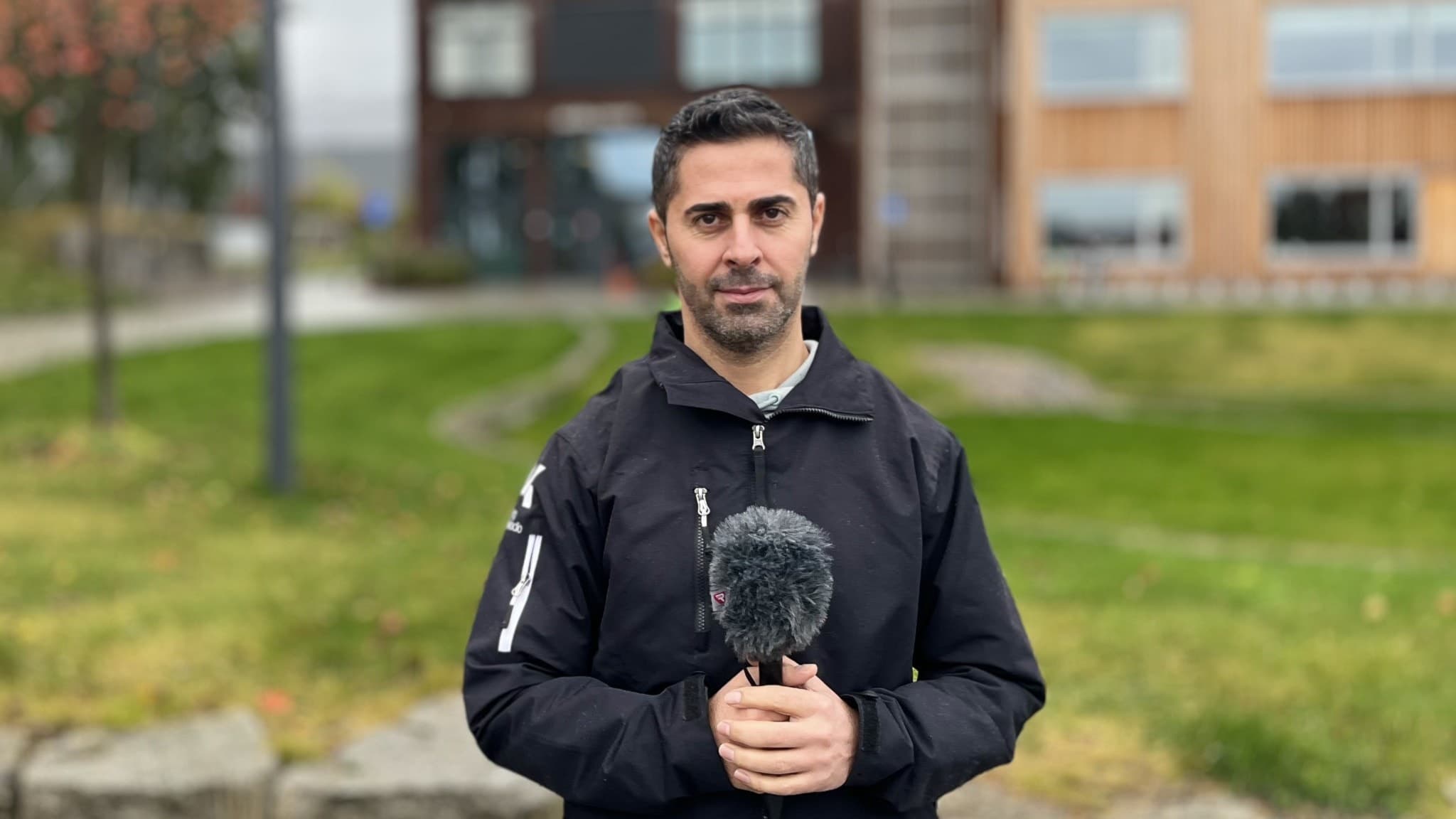 En person står utomhus med en mikrofon i händerna, klädd i en svart jacka. Bakgrunden visar en grön gräsmatta och moderna byggnader.