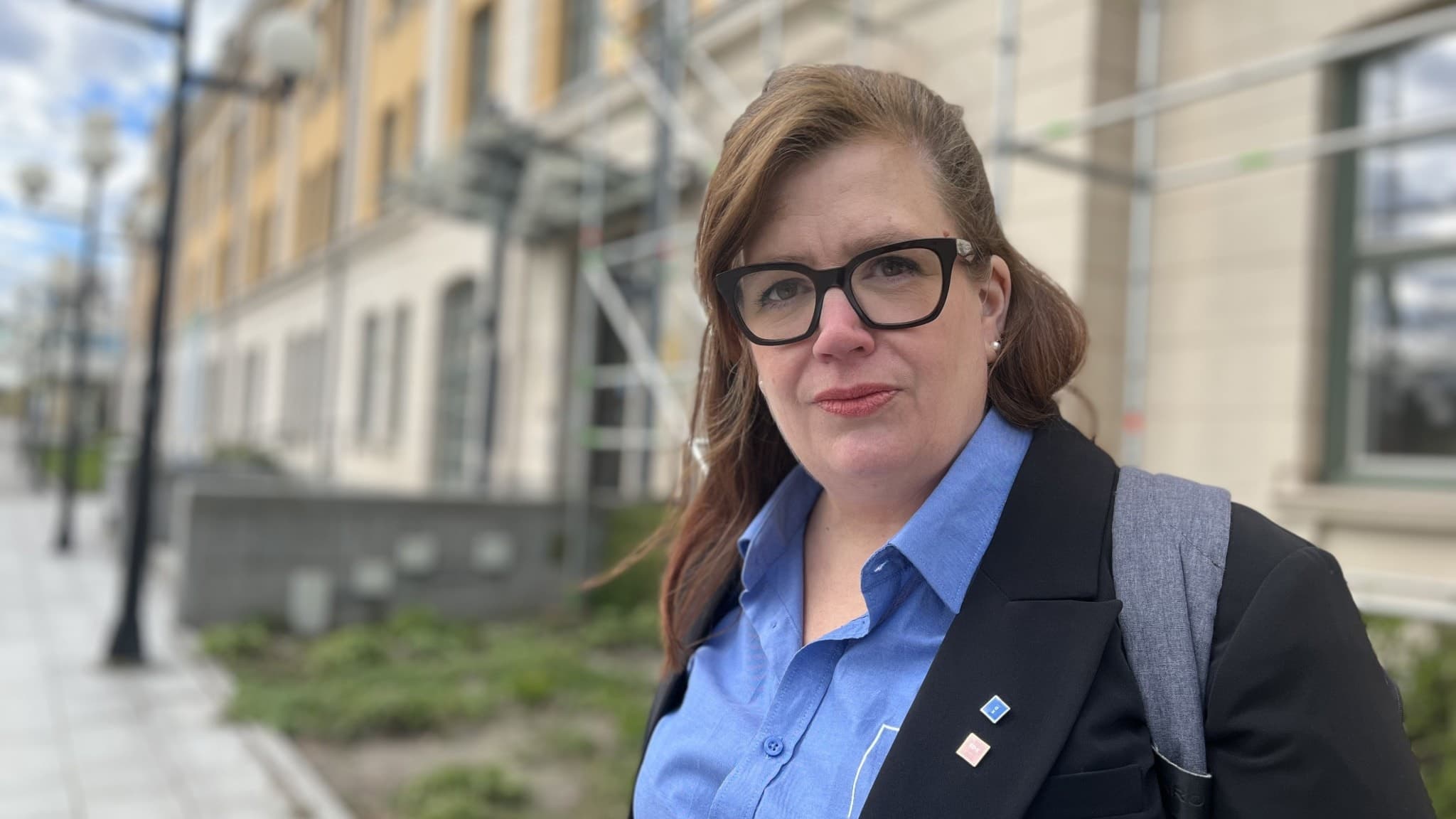 Veronica Kerr står utomhus framför en byggnad med gul och vit fasad, klädd i blå skjorta och mörk kavaj, med en ryggsäck. Bakgrunden visar en trottoar och lyktstolpar.