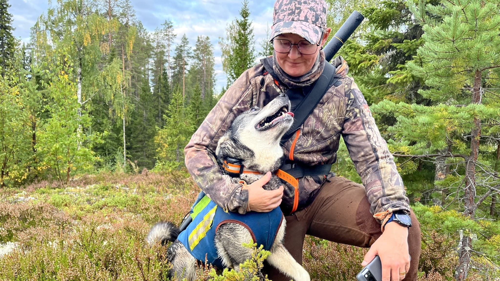 Gerd Lövgren sitter i skogsmiljö med hunden Tikka, som bär en blå väst med gul markering. Gerd har jaktkläder och en bössa över axeln, omgiven av grönskande träd och mossa.