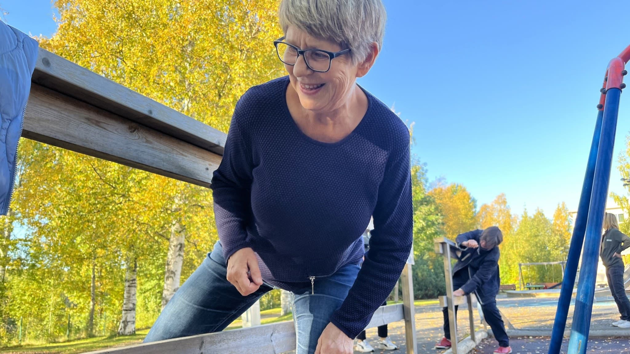 Kvinna med blå tröja utövar seniorparkour genom att huka och ta sig igenom ett staket. I bakgrunden träd med gula höstlöv.
