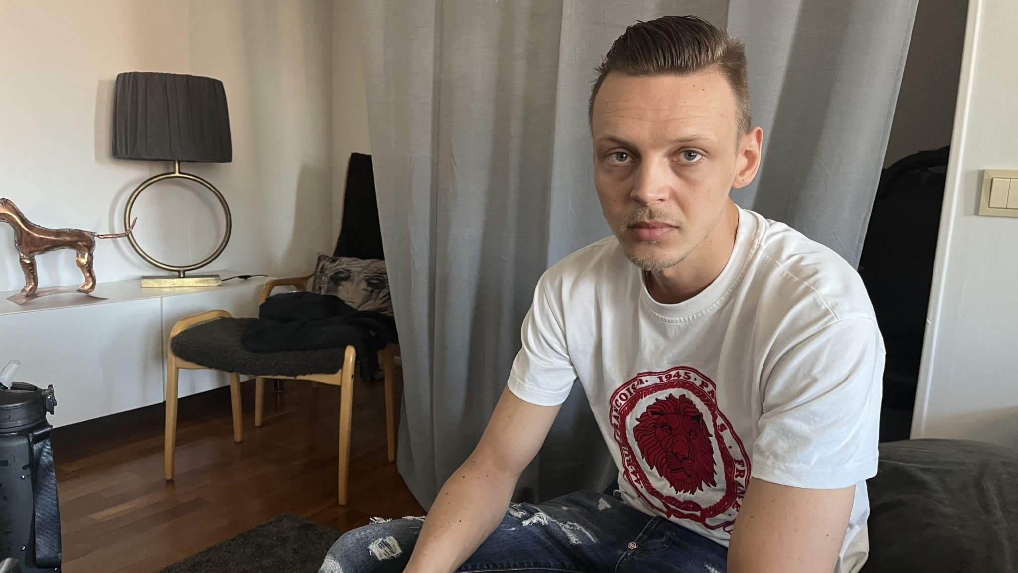 Zebastian sitter på en soffa i ett vardagsrum med grå gardiner, en trästol och en lampa i bakgrunden. Han bär en vit t-shirt med ett rött tryck och jeans.