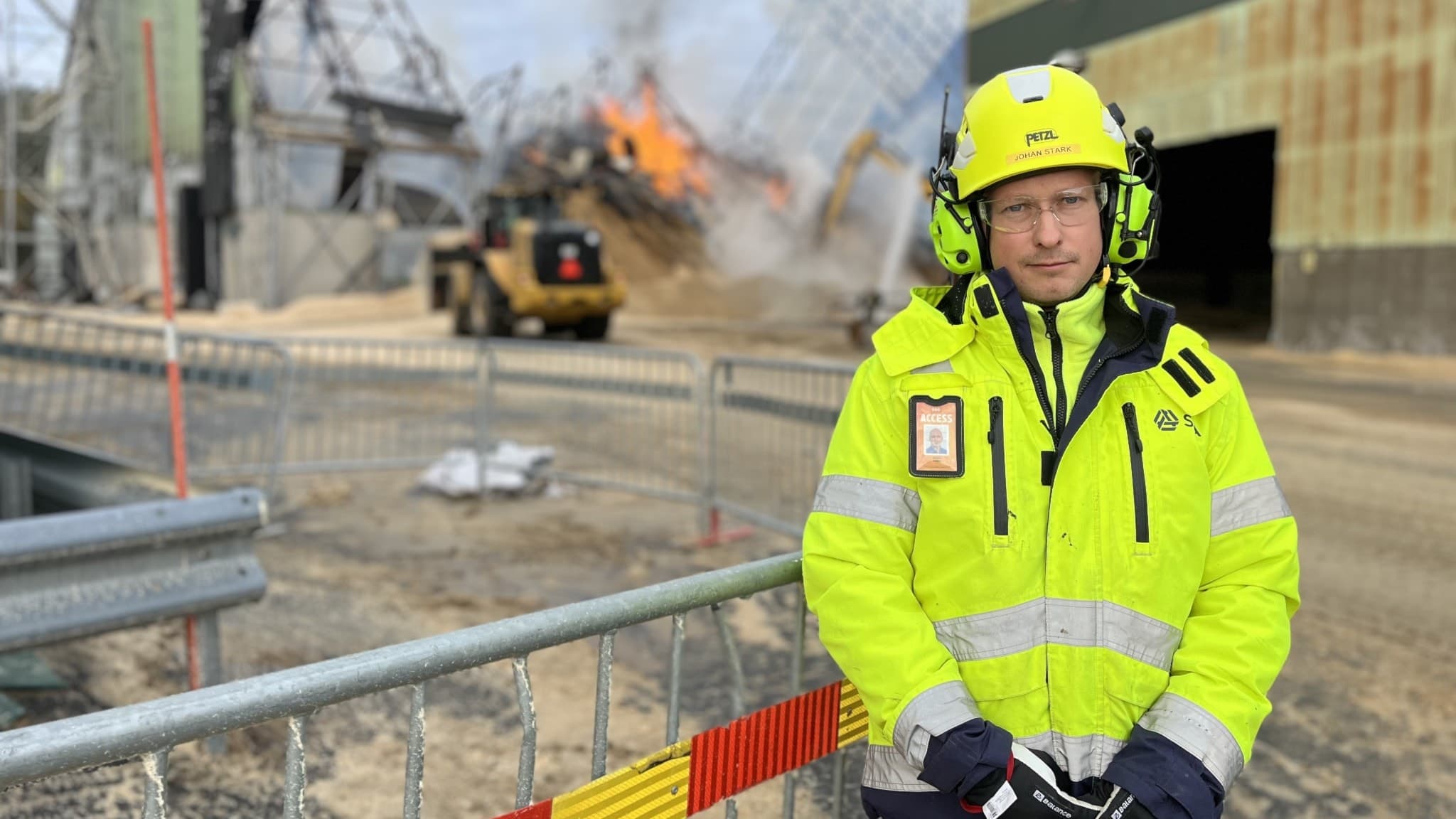 Johan Stark, driftchef och brandansvarig på Bionorr, står i gul skyddsutrustning framför avspärrningar vid en industribyggnad med pågående brand och rökutveckling i bakgrunden.