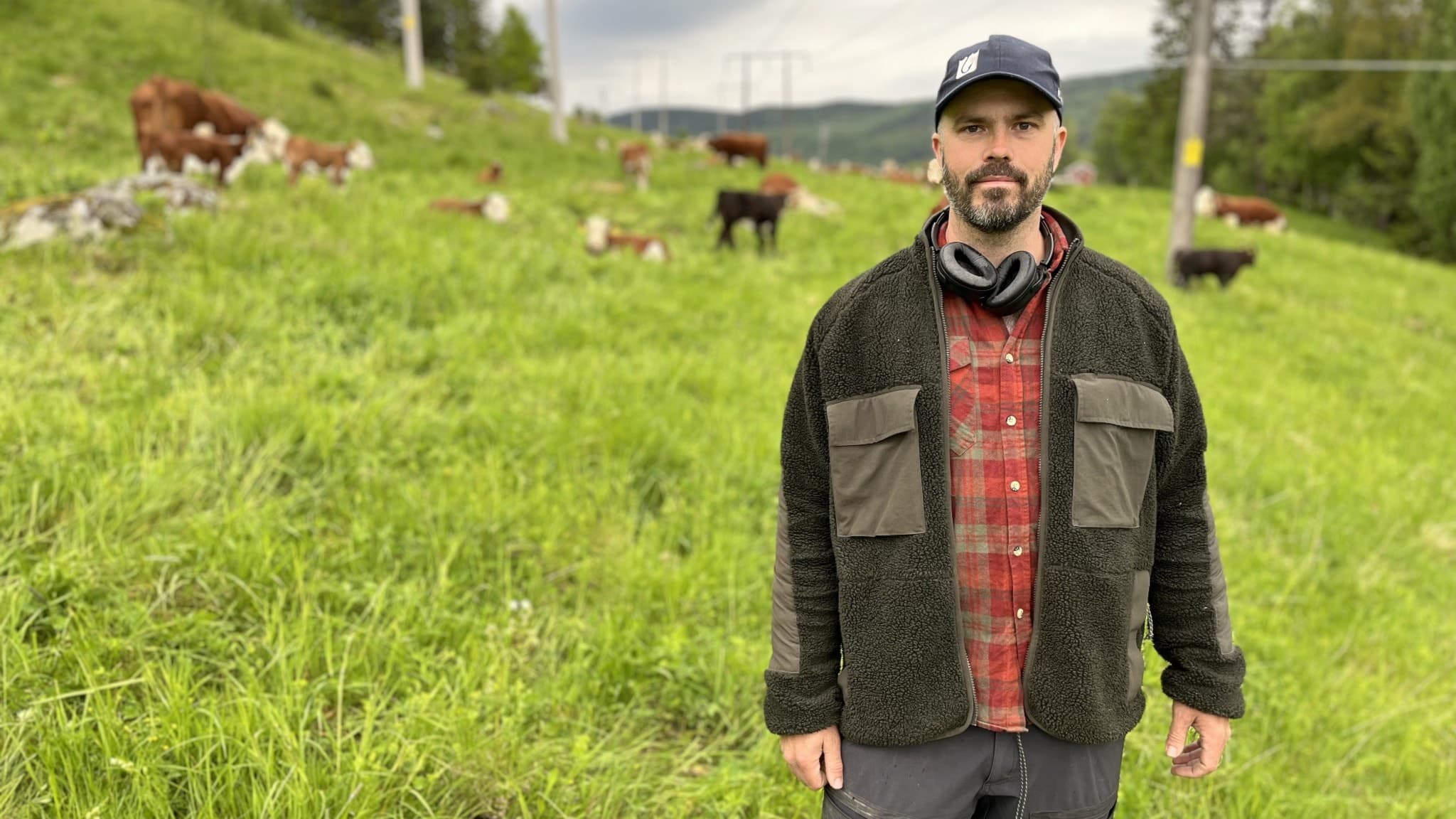 Johan Rudin, ekobonde i Järkvissle, Sundsvalls kommun, står på en grön äng med betande kor i bakgrunden under en molnig himmel. Han har på sig en mörk jacka och röd skjorta.
