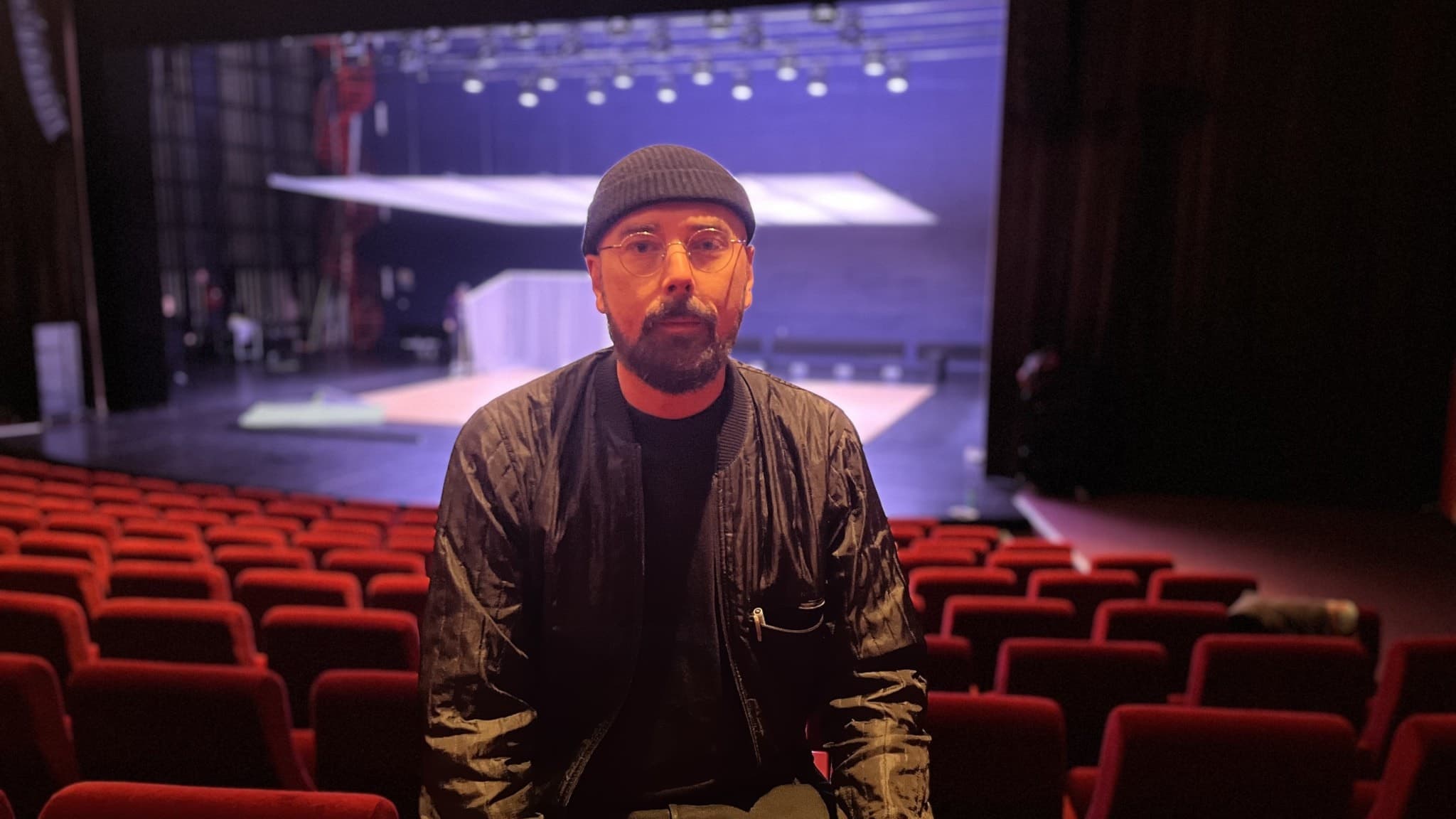 En man med mössa sitter i en teatersalong med ryggen mot scenen. 