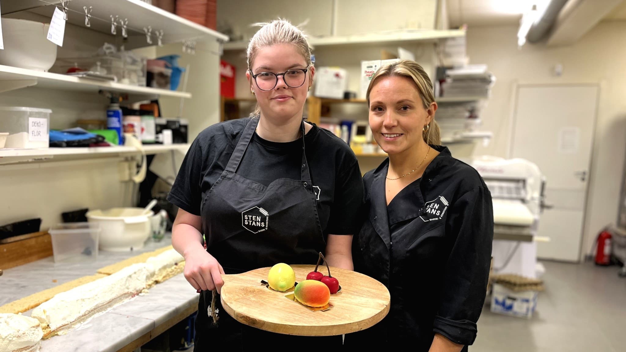 Moa Melander Sandsten, konditor, och Ida Bergafors, ägare av Stenstans Bageri, står i ett bageri iklädda svarta arbetskläder och håller en bricka med bakelser som ser ut som frukter. Bakom dem syns bakredskap och ingredienser.