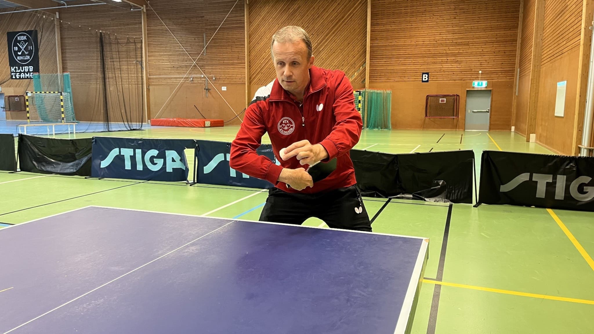 Joakim Carlsson, ordförande i Kungsör Bordtennisklubb, står vid ett bordtennisbord i en idrottshall och håller ett racket. Han är klädd i röd träningsjacka.