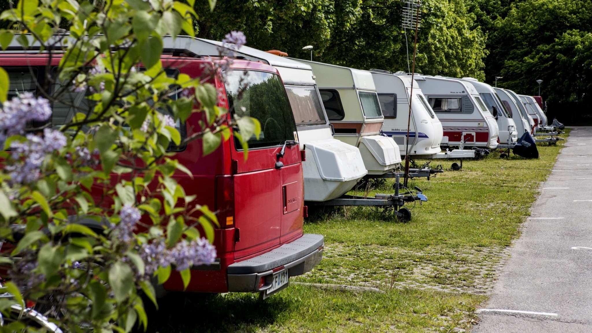 Flera husvagnar står uppställda på en camping, omgivna av grönska och träd. En röd bil syns i förgrunden, och en grusväg löper längs sidan av campingen.