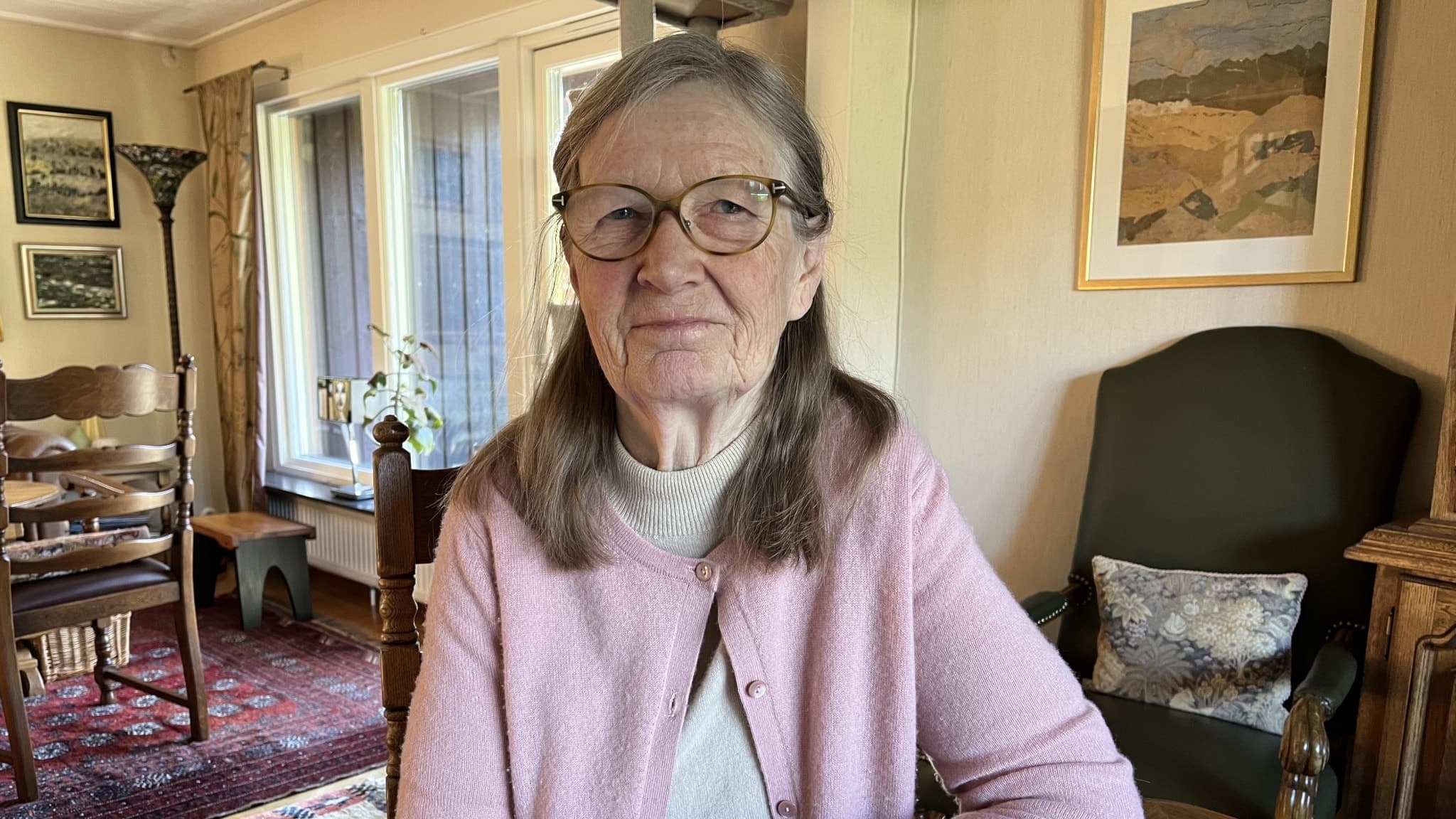 Kerstin Andersson sitter vid ett bord i ett vardagsrum med ljusinsläpp från fönster, omgiven av möbler och tavlor på väggarna. Hon bär en rosa kofta och har grått hår.