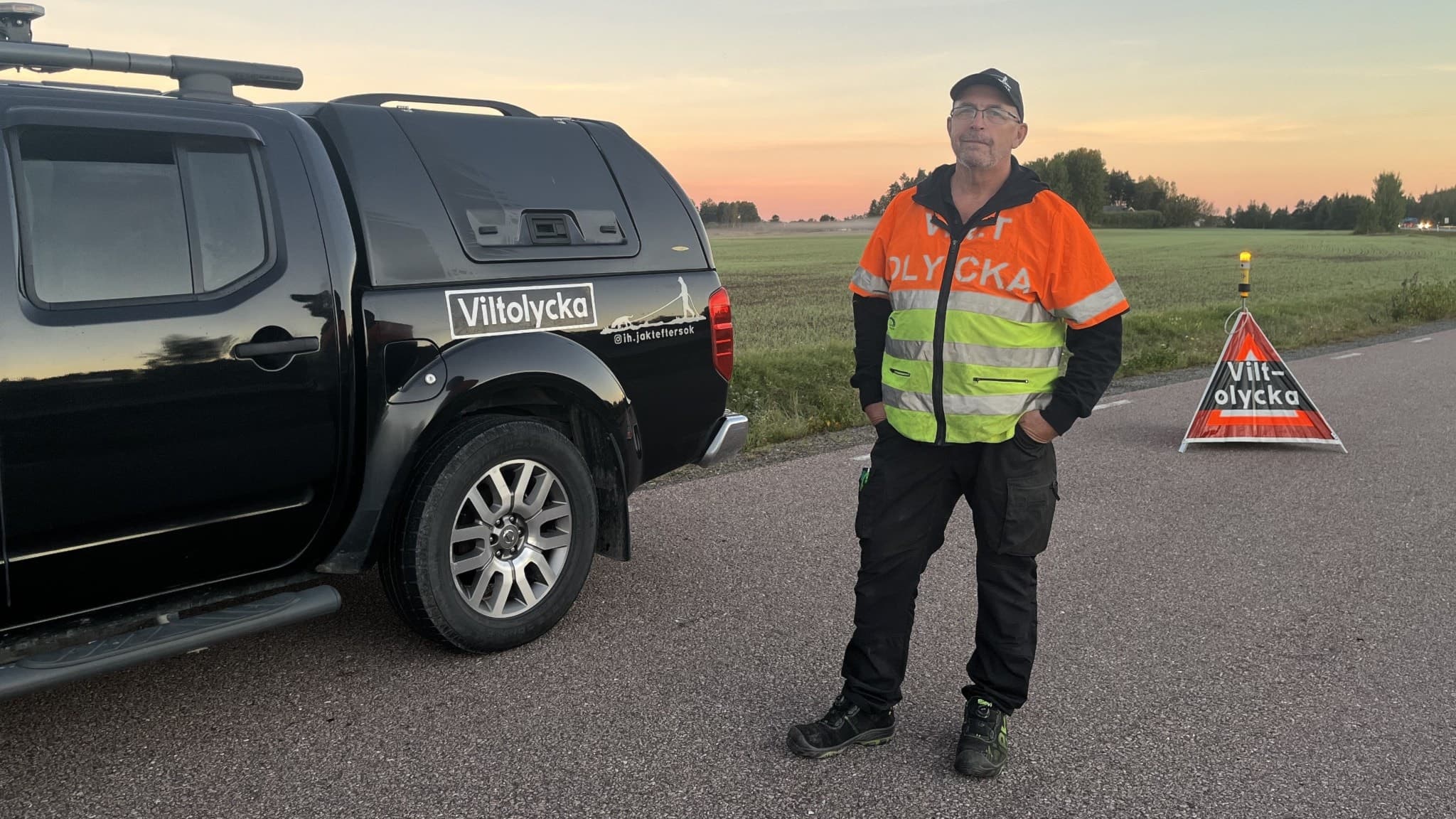 Eftersöksjägaren, Ingvar, står vid en svart bil med jaktutrustning på en landsväg. Han bär en reflexväst och bakom honom syns en varningsskylt samt öppet landskap i solnedgång.