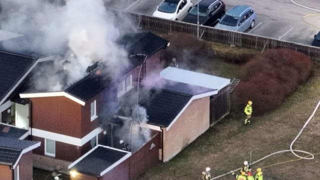 Flygbild över en villa i Skälby, Västerås, där kraftig rök och eld syns efter en misstänkt mordbrand. Brandmän arbetar på plats och flera bilar står parkerade i närheten.