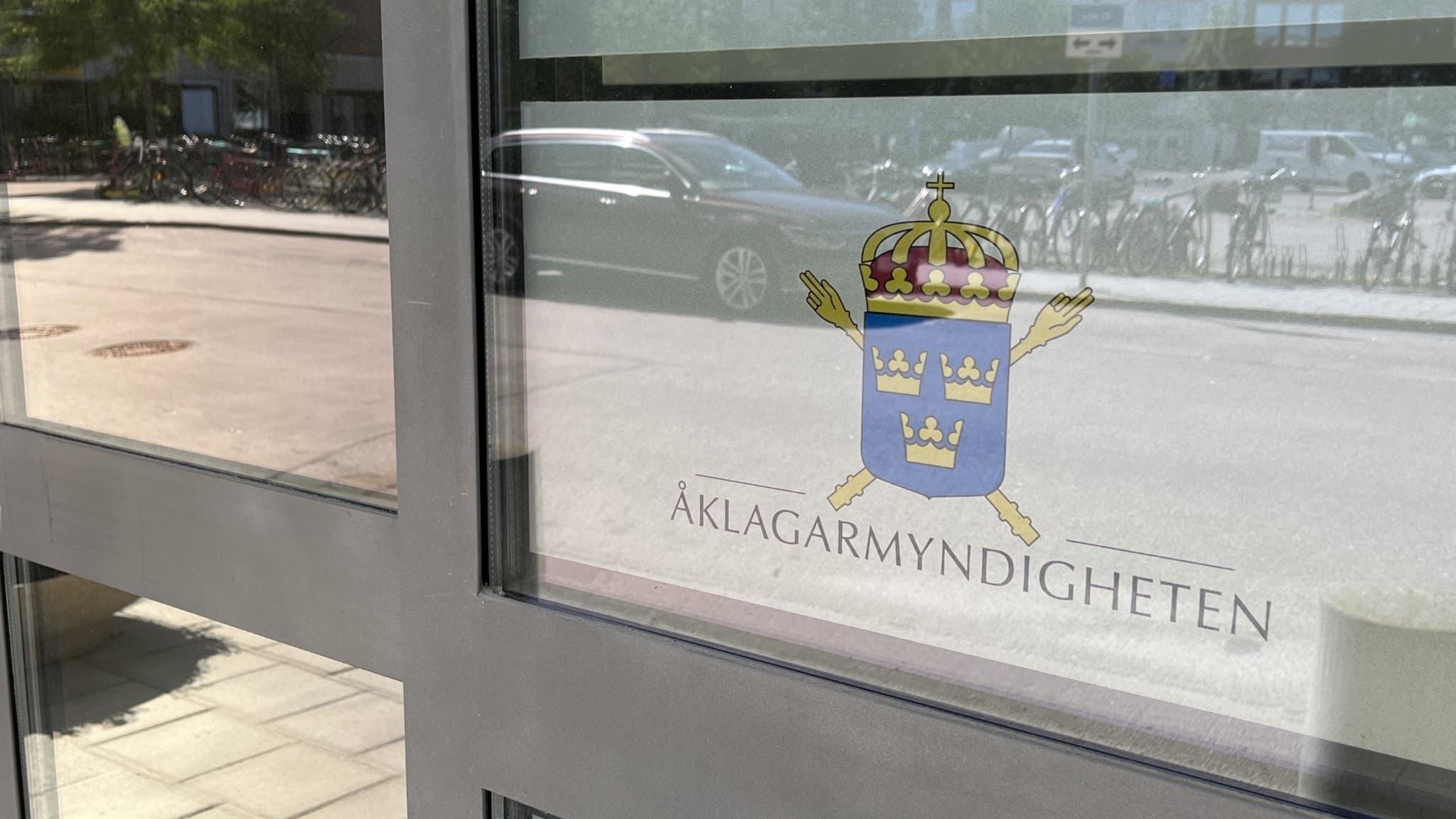 Entrédörr med Åklagarmyndighetens logotyp. Glaset speglar en gata med bilar och cyklar i bakgrunden.