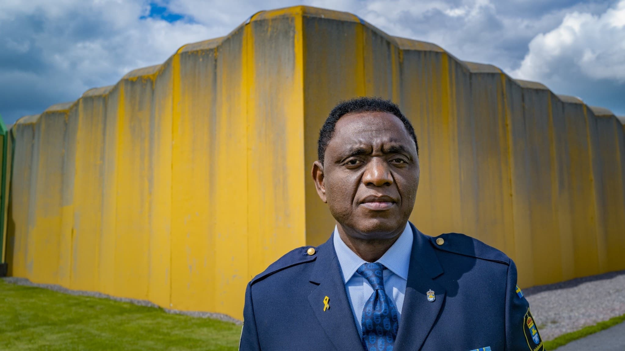 Jacques Mwepu, chef för Kumlaanstalten, står framför en gul mur klädd i en mörkblå uniform med slips, under en molnig himmel.