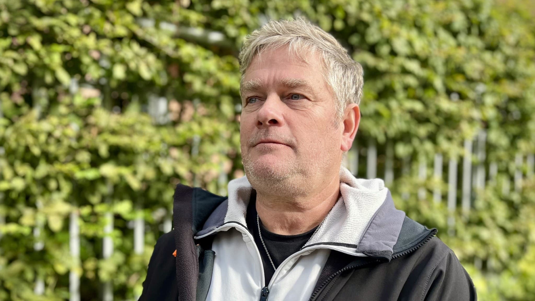 Mikael Eriksson står utomhus framför gröna buskar och ett staket. Han är klädd i en svart jacka och vit tröja med dragkedja.