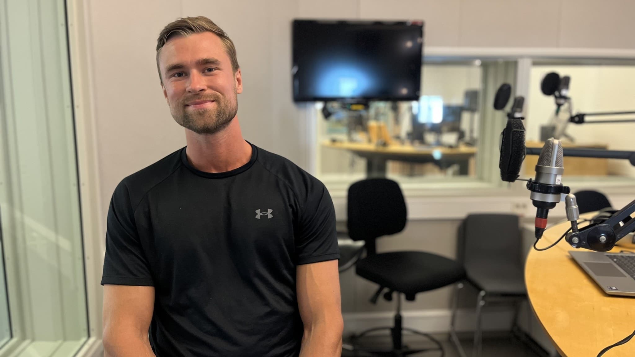 En man sitter i en radiostudio med mikrofoner och skärmar i bakgrunden. Han har på sig en svart träningströja och omgivningen består av modern studioutrustning.