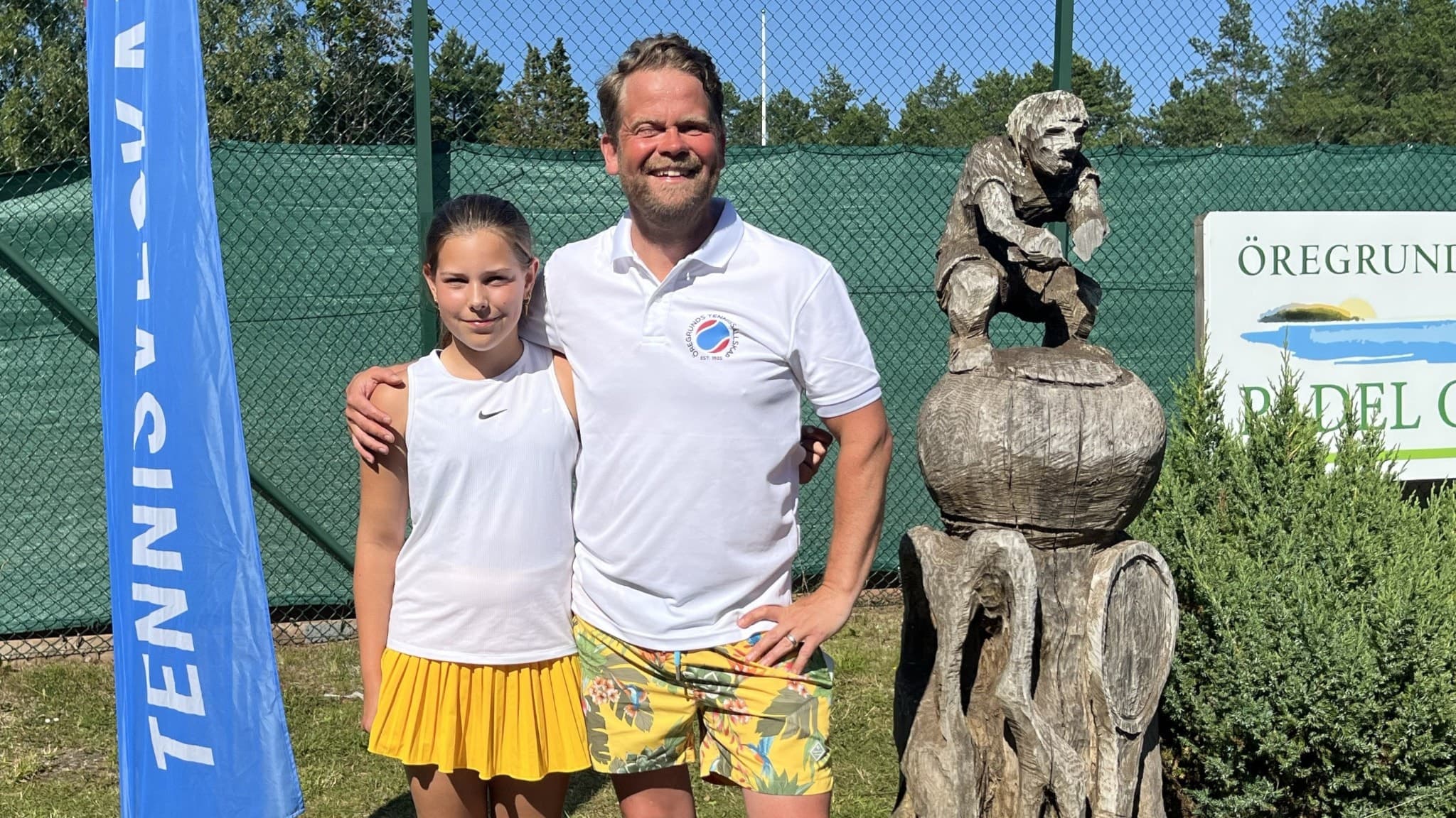 Malla Strömgren och Andreas Pragsten står utomhus, klädda i sportkläder. En dekorativ träskulptur och en skylt med texten 'Öregrund' syns i bakgrunden.