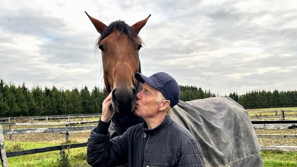 Stefan Melander står bredvid en brun häst med täcke på en hästgård, omgiven av staket och en skog i bakgrunden under en molnig himmel.