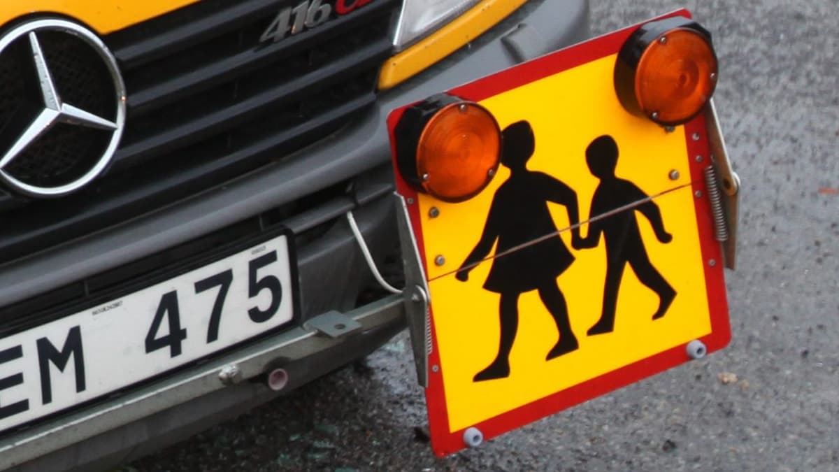 Närbild på en skolskjuts med ett varningsskylt som visar två barn som går hand i hand. Skylten är fäst på fronten av en Mercedes-buss med registreringsnummer EM 475.