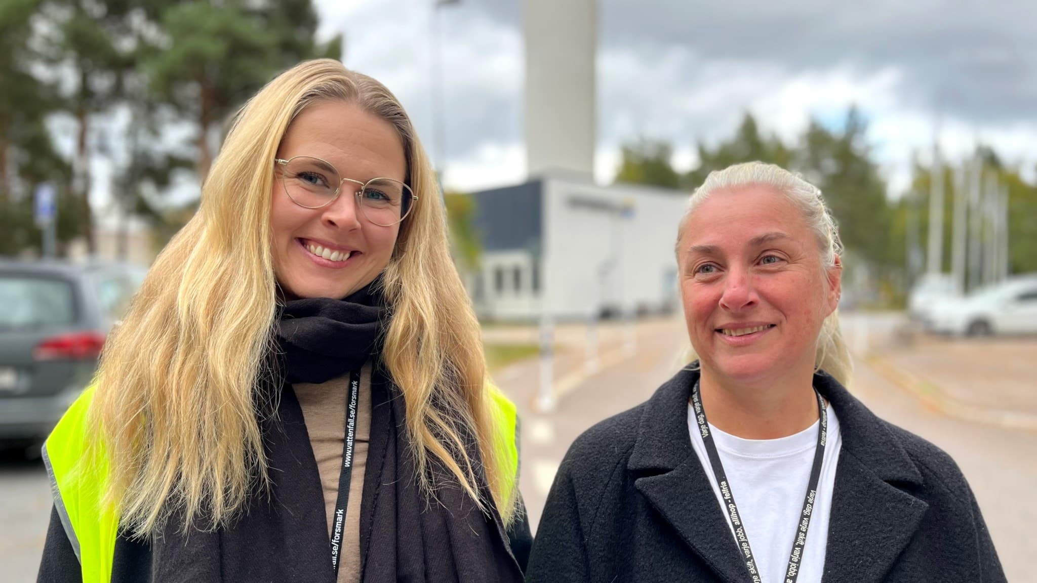Jennifer och Jessica, klädda i arbetskläder, står utomhus och ler framför Forsmarks kärnkraftverk. De medverkar i tv-serien "Högspänning".