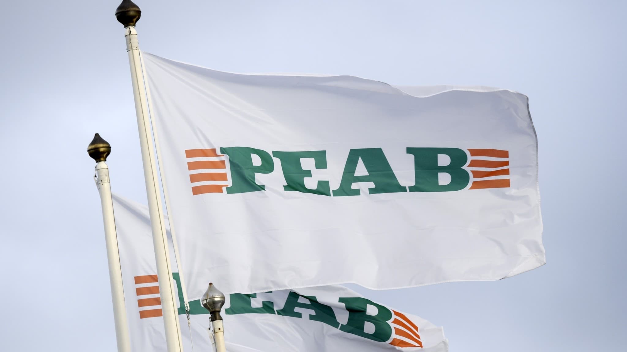 Fladdrande flaggor med byggföretaget PEABs logotyp mot en ljus himmel. Logotypen består av gröna bokstäver med orangea linjer vid sidorna.