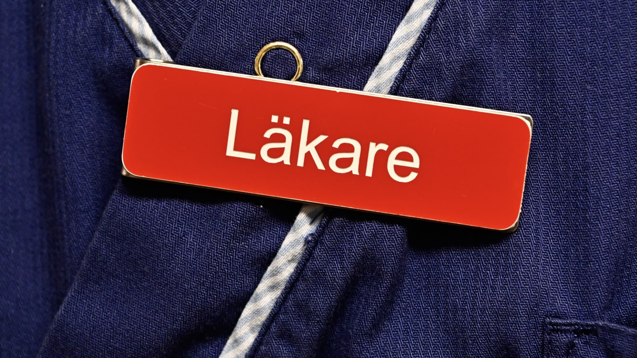 Närbild på en röd namnbricka med texten 'Läkare', fäst på en blå läkarrock. 