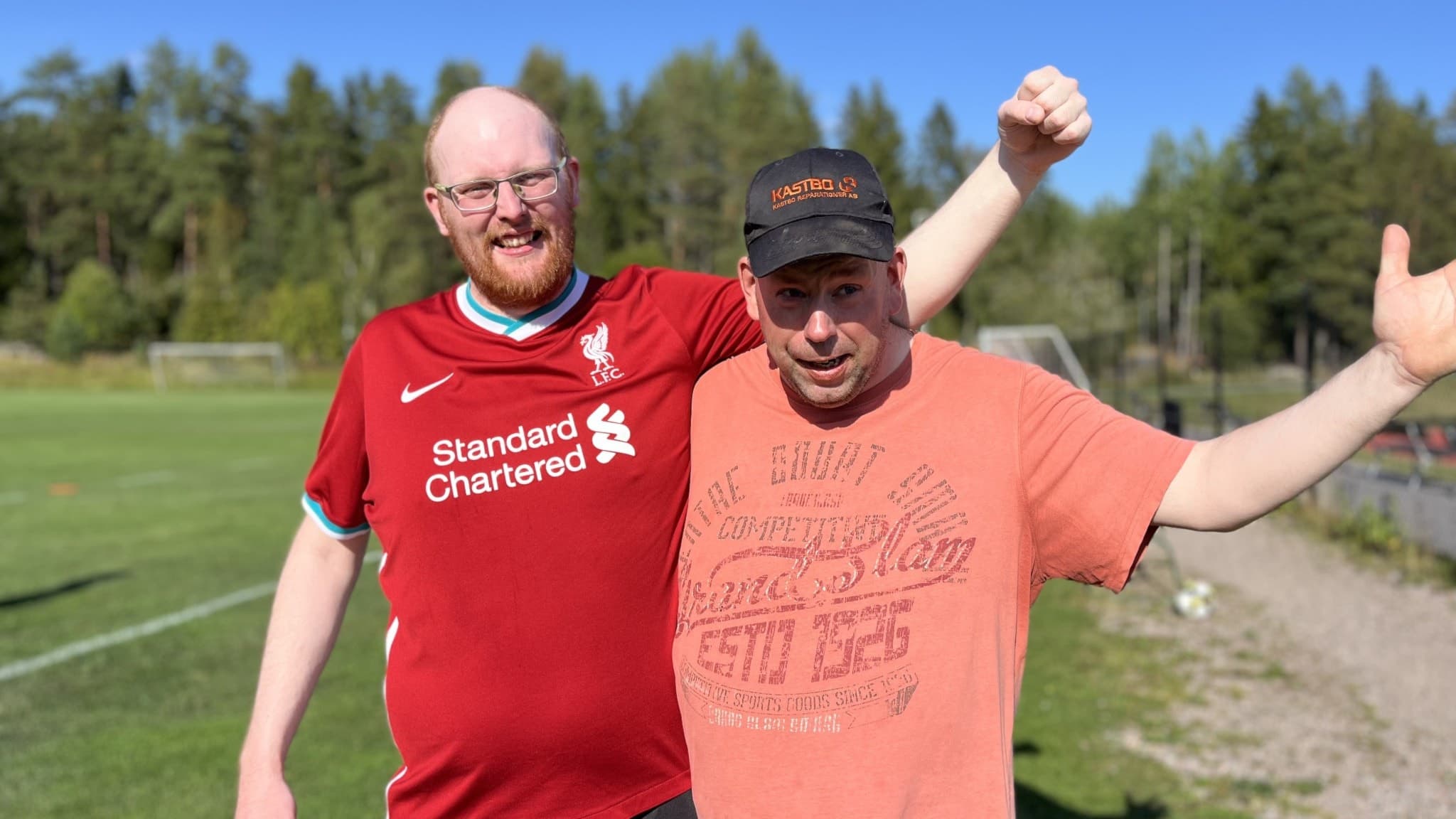 Två personer står på en gräsplan och visar glädje. Den ena bär en röd fotbollströja och den andra en orange t-shirt och keps. Skog syns i bakgrunden under en klarblå himmel.
