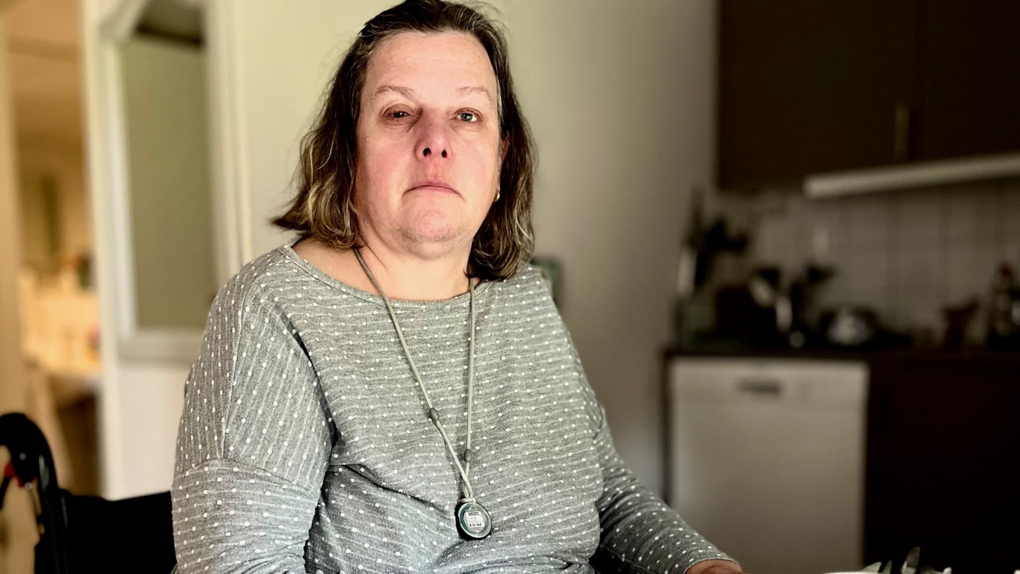 En kvinna i rullstol sitter vid ett köksbord med en kopp i förgrunden, iklädd en grå tröja. Bakom henne syns ett kök med diskbänk och skåp i en hemmiljö.