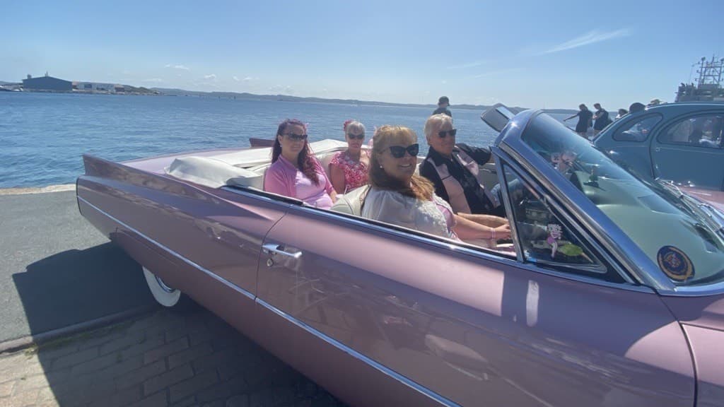 Fyra personer sitter i en rosa veteranbil vid en kaj med havet och blå himmel i bakgrunden. Scenen förmedlar en känsla av sommar och retrostil.