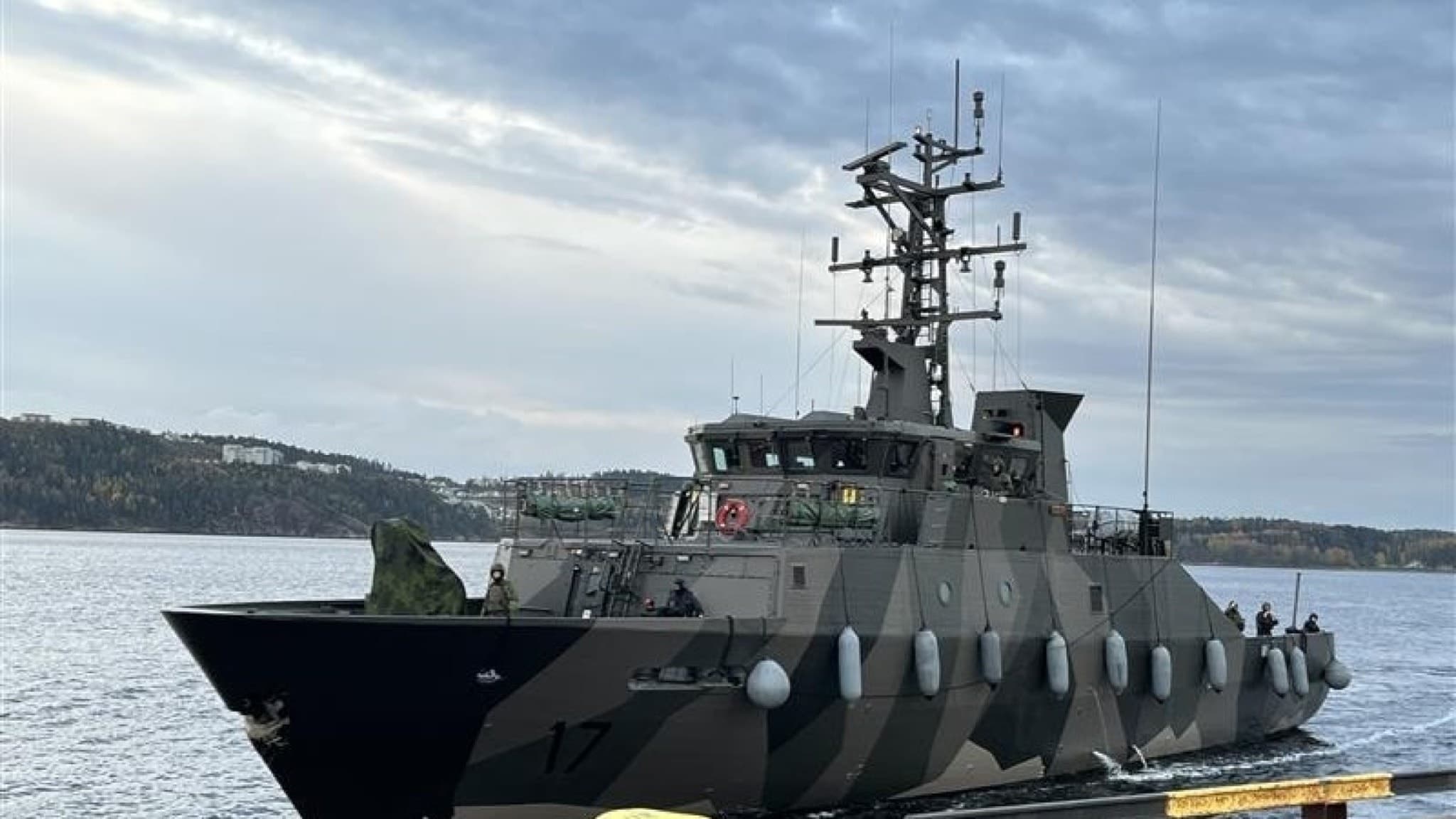 Militärbåt i kamouflagefärger seglar på vattnet nära Uddevalla, med skogsklädda kullar i bakgrunden under en molnig himmel.