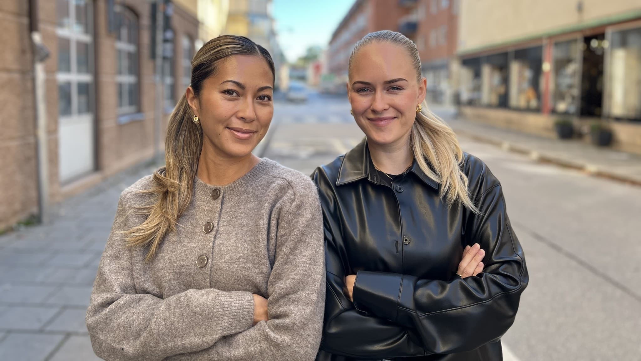 Hanna Lauste och Linn Rosenqvist står med armarna i kors på en gata omgivna av byggnader. De har avslappnade ansiktsuttryck och är klädda i höstkläder.