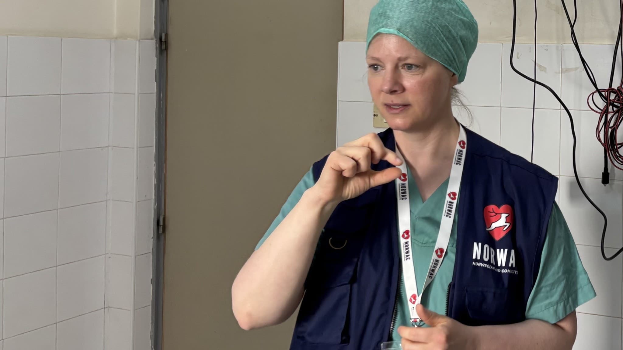 Operationssjuksköterskan Nina Maria Grönn Hvinden står i ett sjukhusmiljö och bär en mörkblå väst med Norwac-logotypen, gröna sjukhuskläder och en ljusgrön operationsmössa.