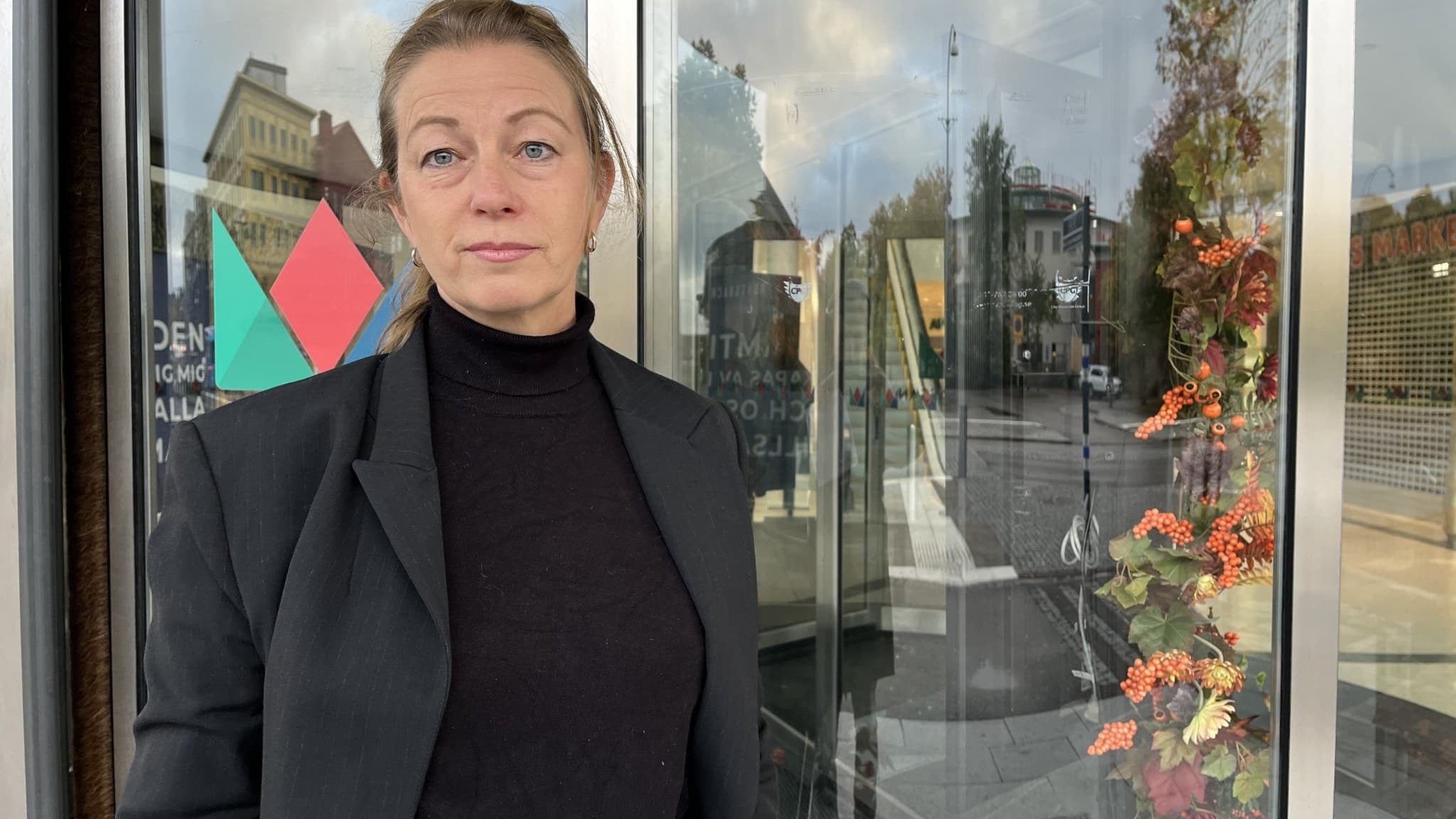 En person står framför en glasdörr till en byggnad, klädd i svart kavaj och polotröja. Höstdekorationer syns på dörren och en stadsmiljö med byggnader och träd i bakgrunden.